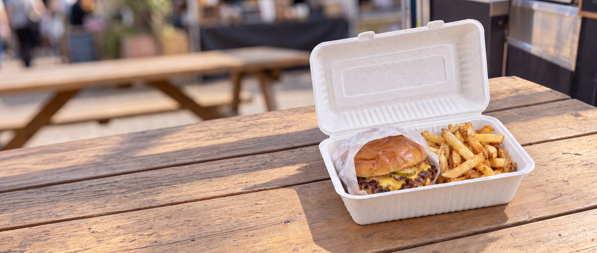 Smash burger and fries in an open white bagasse 9x6 clamshell on a wooden table at an outdoor street food market